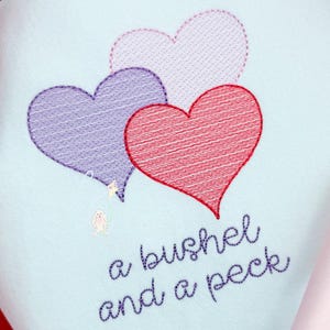 May include: Embroidered design featuring three hearts in shades of purple, red, and pink on a light blue background. The text "a bushel and a peck" is stitched below the hearts. A small bunny and stars are also included.