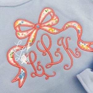 May include: Light blue sweatshirt with a coral-colored embroidered bow and the initials "LK" in a script font. The bow features a floral pattern in pastel colors. A small bunny illustration is also present.