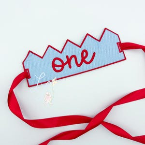 May include: A blue and white gingham crown with red trim and the word "one" in red script. The crown has a red ribbon attached. A small bunny with a balloon is attached to the crown.