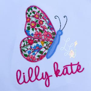 May include: White fabric with a large embroidered butterfly. The butterfly has a blue body and antennae, and its wings are patterned with pink, red, blue, and yellow flowers. The name "lilly kate" is embroidered in pink script below the butterfly.