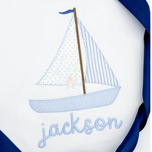 May include: White fabric with a blue sailboat embroidery. The sailboat has a blue hull, a striped sail, and a dotted sail. The name "jackson" is embroidered in blue script below the boat. A blue ribbon is in the background.