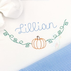 May include: A white fabric garment featuring an embroidered design of a pumpkin with green vines and the name Lillian in blue script. A large white fabric bow is attached at the top left, and a blue and white gingham fabric panel is at the bottom.