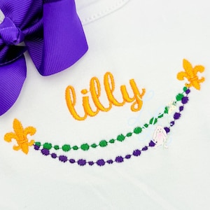 May include: White garment with the name "Lilly" embroidered in orange script. The design includes green and purple beaded strands, gold fleur-de-lis accents, and a small pink bunny. A large purple bow is visible.