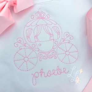 May include: White garment featuring a pink embroidered design of a fairytale carriage. The carriage has detailed wheels and a decorative top. The name "phoebe" is embroidered in cursive below the carriage. A pink bow and ribbon are in the background.