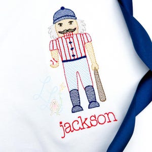 Könnte beinhalten: Weißer Stoff mit einer Stickerei eines Baseball-Nussknackers. Der Nussknacker trägt eine blaue Mütze, ein rot-weiß gestreiftes Hemd und eine blaue Hose. Der Name "jackson" ist darunter in roter Schrift gestickt.