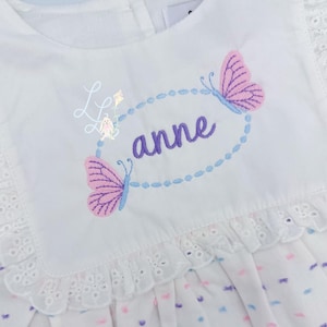 May include: White dress with embroidered butterflies and the name "anne" in purple. The dress features a ruffled collar and hem with eyelet lace detailing. The design includes a kite and a bunny.
