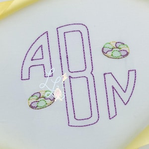 May include: White fabric with a purple embroidered monogram of the letters ABN. Two small, embroidered circular designs with green, purple, and yellow accents are also present. A yellow ribbon is in the upper left corner.