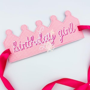 Può includere: Una corona in tessuto a quadretti rosa con le parole "birthday girl" ricamate in rosa. La corona ha un nastro di raso rosa attaccato per l'allacciatura. Un piccolo coniglietto e un ornamento a stella sono sulla corona.