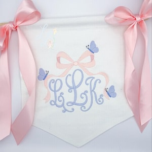 May include: White banner with pink satin ribbons and a wooden dowel. The banner features a light blue monogram with the letters LLK, surrounded by a pink bow and three light blue butterflies. A small illustration of a bunny holding a balloon is also present.