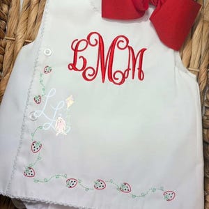May include: White baby romper with red embroidered initials and a red bow. The romper features a strawberry vine design and a small illustration of a bunny with a kite. The text "Stitched by PK & Co Embroidery" is at the bottom.