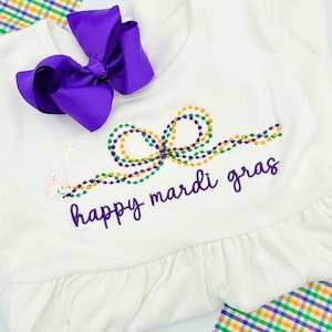 May include: A white baby outfit with a purple bow and the embroidered phrase "happy mardi gras." The design includes a string of beads in green, gold, and purple, along with a small illustration of a bunny holding a kite. The outfit has a ruffled hem.