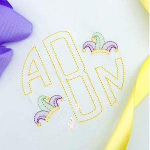 Puede incluir: Una tela blanca con un monograma bordado en oro de las letras ABN dentro de un círculo. El diseño incluye pequeños elementos caprichosos en tonos morados, verdes y amarillos, con un conejito y una estrella. Un lazo morado y una cinta amarilla están en las esquinas.