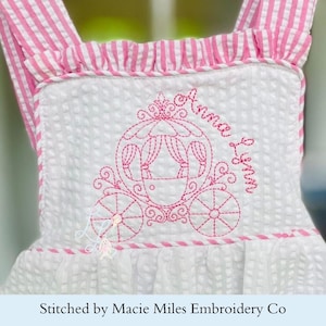 May include: White seersucker bib with pink and white striped straps and trim. The bib features a pink embroidered carriage design with the name "Anna Lynn" in cursive. The bib is part of a baby or toddler clothing set.