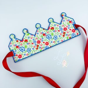 May include: A floral fabric crown with a blue trim and red ribbon. The crown features a pattern of red strawberries, blue and red flowers, and green leaves. The red ribbon is attached to the crown's sides.