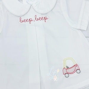 Könnte beinhalten: Weißes kurzärmeliges Hemd mit einem Bubikragen. Das Hemd hat einen roten gestickten Text mit der Aufschrift "beep, beep" und ein kleines gesticktes Auto. Ein kleines gesticktes Design eines Hasen und eines Drachens ist ebenfalls vorhanden.