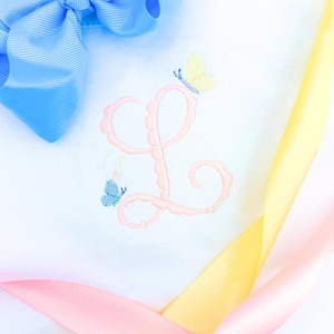 Puede incluir: Tela blanca con una letra "L" bordada en rosa claro y dos mariposas bordadas, una amarilla y otra azul. Un lazo azul claro está en la esquina superior izquierda, y cintas rosas y amarillas en la esquina inferior derecha.