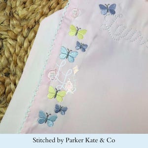 May include: Pale pink embroidered fabric with butterflies in shades of blue and yellow, the name "Laya" in cursive, and a bunny with a kite. The fabric has a scalloped edge and button details. The text "Stitched by Parker Kate & Co" is at the bottom.