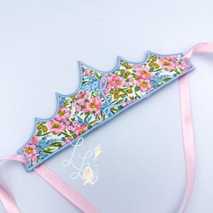 May include: A floral fabric crown with a scalloped edge, featuring pink and blue flowers on a white background. The crown is outlined in light blue stitching and has pink satin ribbons attached. The letter "L" is embroidered on the crown.