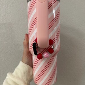 May include: A pink and white striped tumbler with a pink handle. A red and pink beaded wristlet is attached to the handle. The tumbler has a clear lid and a red rim.