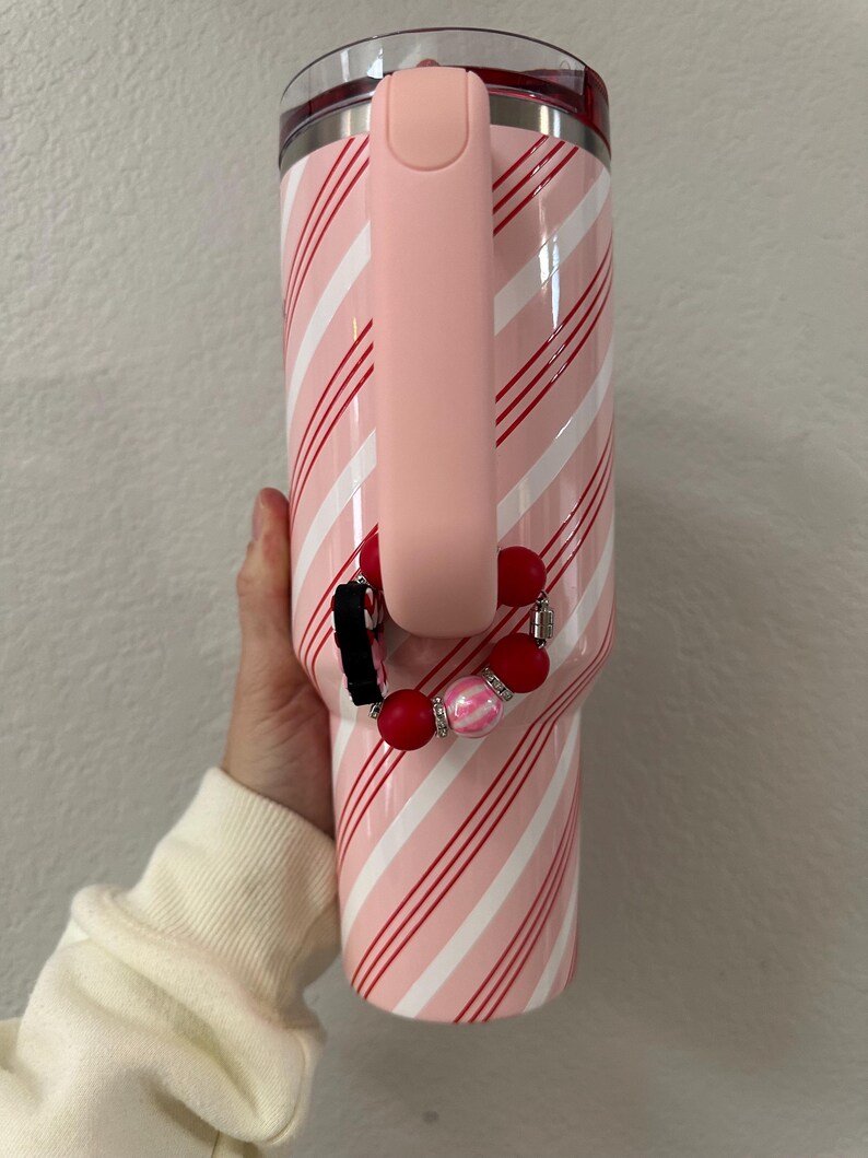 May include: A pink and white striped tumbler with a pink handle. A red and pink beaded wristlet is attached to the handle. The tumbler has a clear lid and a red rim.