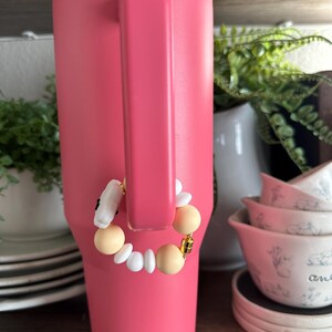 May include: A large, pink insulated tumbler with a handle. A decorative charm bracelet with white, beige, and gold beads is attached to the handle. The tumbler is displayed on a wooden shelf with other kitchenware.