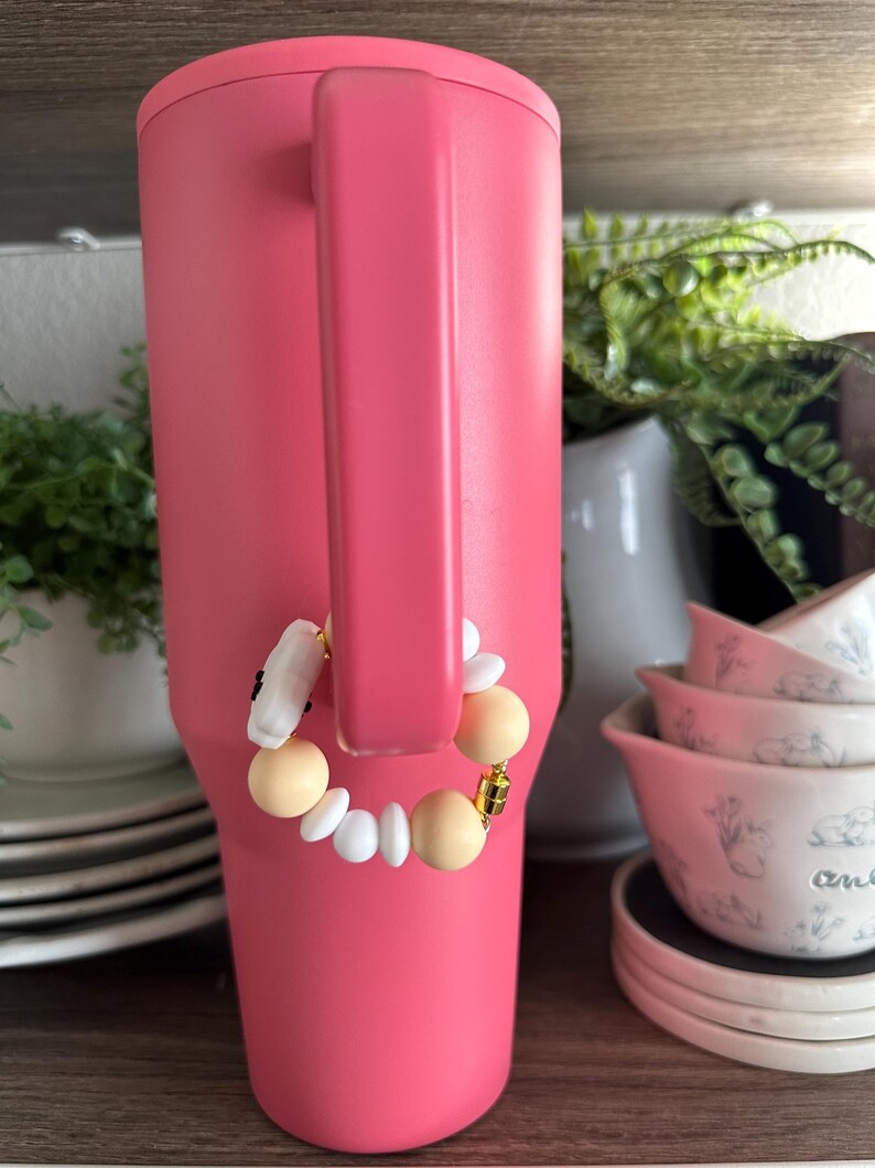 May include: A large, pink insulated tumbler with a handle. A decorative charm bracelet with white, beige, and gold beads is attached to the handle. The tumbler is displayed on a wooden shelf with other kitchenware.