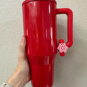 Puede incluir: Un vaso t&eacute;rmico rojo brillante con asa y tapa a juego. Un adorno de copo de nieve rosa est&aacute; sujeto al asa. El vaso est&aacute; sostenido por una persona.