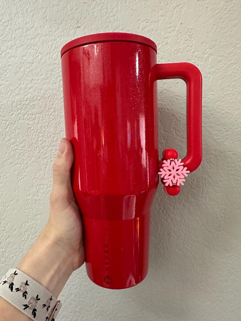 Puede incluir: Un vaso t&eacute;rmico rojo brillante con asa y tapa a juego. Un adorno de copo de nieve rosa est&aacute; sujeto al asa. El vaso est&aacute; sostenido por una persona.