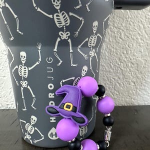 Puede incluir: Un vaso con tem&aacute;tica de Halloween con un asa negra y un patr&oacute;n de esqueletos blancos. Una pulsera de cuentas moradas y negras con un dije de sombrero de bruja est&aacute; en primer plano. El vaso tiene el texto "HYDROJUG" y "40oz".