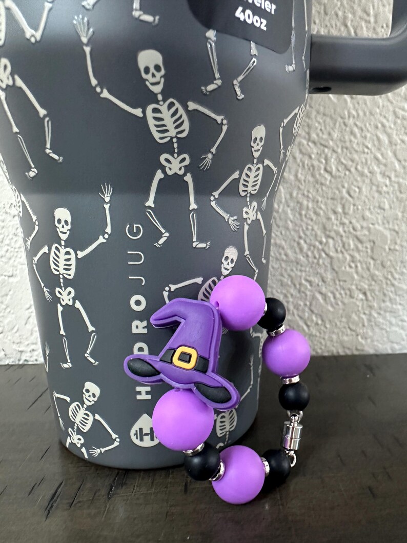 Puede incluir: Un vaso con tem&aacute;tica de Halloween con un asa negra y un patr&oacute;n de esqueletos blancos. Una pulsera de cuentas moradas y negras con un dije de sombrero de bruja est&aacute; en primer plano. El vaso tiene el texto "HYDROJUG" y "40oz".