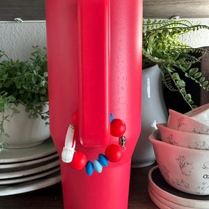 May include: A vibrant red insulated tumbler with a matching handle. A decorative charm with red, white, and blue beads is attached to the handle. The tumbler is displayed on a wooden shelf, with other kitchenware in the background.