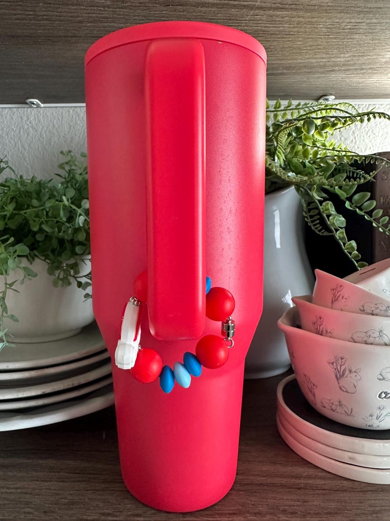 May include: A vibrant red insulated tumbler with a matching handle. A decorative charm with red, white, and blue beads is attached to the handle. The tumbler is displayed on a wooden shelf, with other kitchenware in the background.