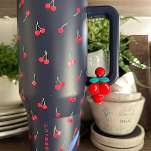 May include: A navy blue HydroJug water bottle with a handle, covered in a pattern of red cherries. The word "HYDROJUG" is printed vertically on the side. A cherry charm hangs from the handle. The bottle is approximately 10 inches tall.