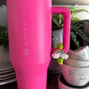 May include: A bright pink HydroJug water bottle with a handle. The bottle has the HydroJug logo printed on the side. A charm of a rainbow and clouds is attached to the handle. The bottle is next to a stack of white bowls.