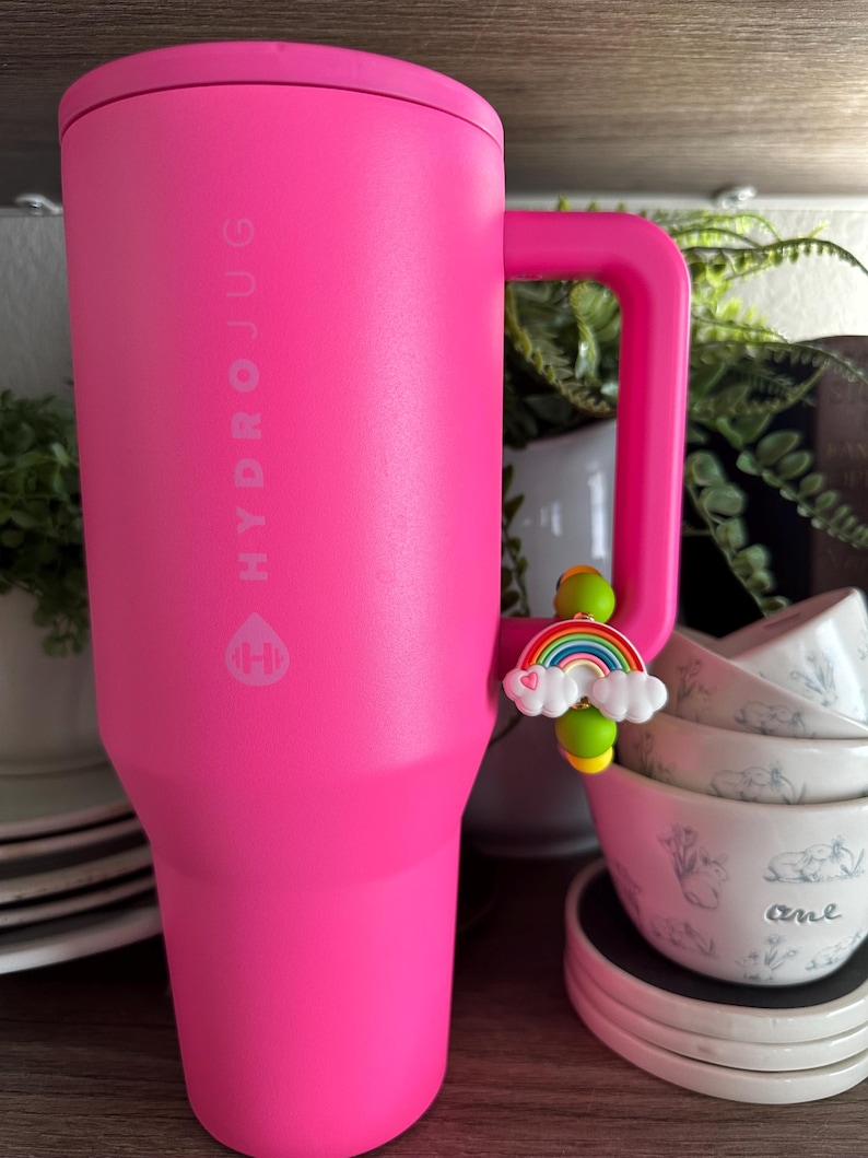 May include: A bright pink HydroJug water bottle with a handle. The bottle has the HydroJug logo printed on the side. A charm of a rainbow and clouds is attached to the handle. The bottle is next to a stack of white bowls.