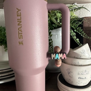 Puede incluir: Un vaso Stanley rosa con borde y asa dorados. La palabra "STANLEY" est&aacute; impresa en el lateral. Un dije que dice "MAMA" en letras coloridas est&aacute; unido al asa. El vaso est&aacute; junto a cuencos blancos con un dise&ntilde;o floral.