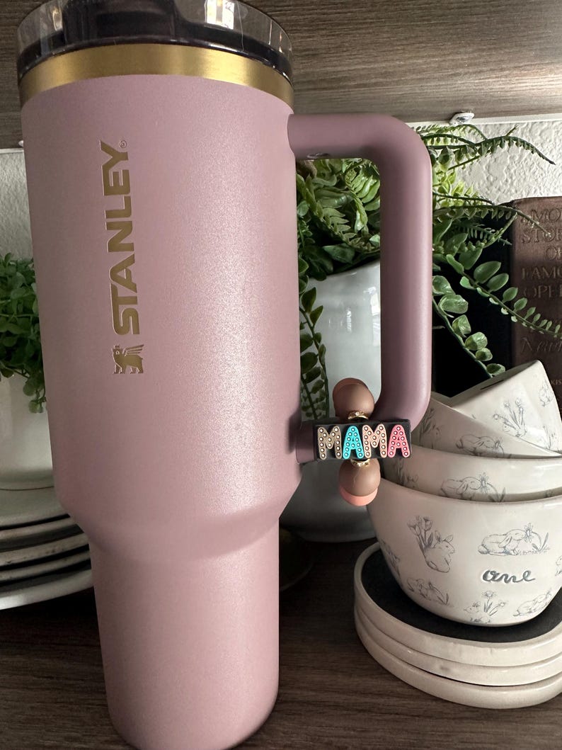Puede incluir: Un vaso Stanley rosa con borde y asa dorados. La palabra "STANLEY" est&aacute; impresa en el lateral. Un dije que dice "MAMA" en letras coloridas est&aacute; unido al asa. El vaso est&aacute; junto a cuencos blancos con un dise&ntilde;o floral.