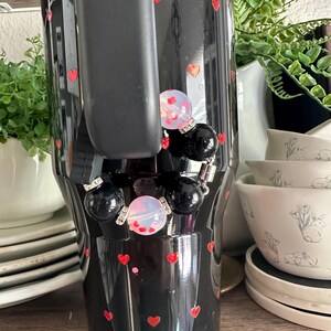 May include: Black tumbler decorated with red hearts. The tumbler has a black handle and is adorned with a charm featuring black and opalescent beads. The background includes white dishes and greenery.
