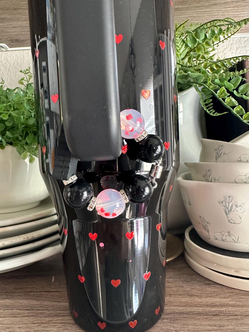 May include: Black tumbler decorated with red hearts. The tumbler has a black handle and is adorned with a charm featuring black and opalescent beads. The background includes white dishes and greenery.