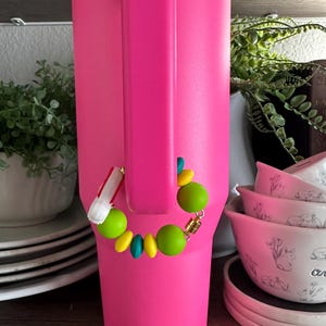May include: A bright pink insulated tumbler with a matching handle. A colorful beaded bracelet with green, yellow, and teal beads is attached to the handle. The background includes white bowls with floral designs and greenery.