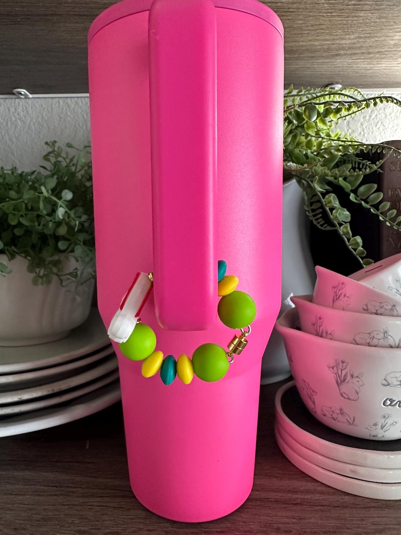 May include: A bright pink insulated tumbler with a matching handle. A colorful beaded bracelet with green, yellow, and teal beads is attached to the handle. The background includes white bowls with floral designs and greenery.