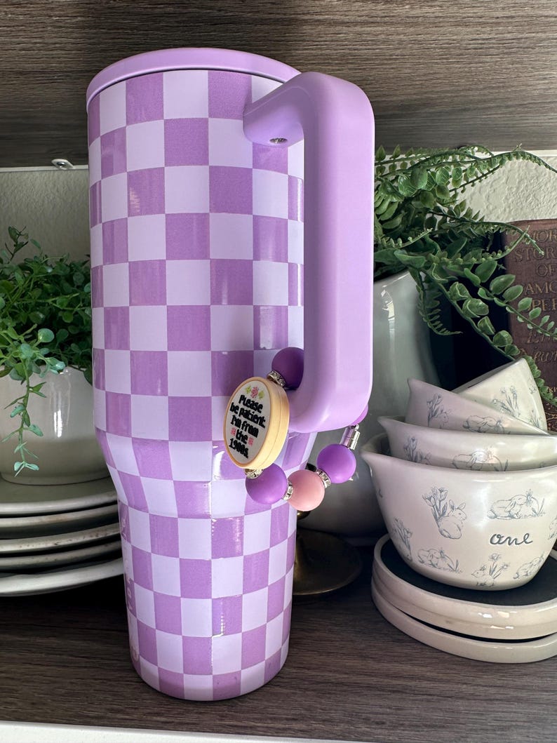 Puede incluir: Un vaso grande y aislado con un patr&oacute;n de cuadros lavanda y un asa a juego. Un dije de cuentas con el texto "Please be patient, I'm from the 1980s" cuelga del asa. El vaso est&aacute; dise&ntilde;ado para bebidas fr&iacute;as.