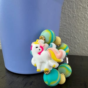 K&ouml;nnte beinhalten: Ein wei&szlig;er Einhorn-Anh&auml;nger mit rosa, gelben und blauen Akzenten ist an einem Perlenarmband befestigt. Das Armband hat bunte, marmorierte Perlen in Blau-, Gelb- und Gr&uuml;nt&ouml;nen. Der Anh&auml;nger und das Armband sind vor einem hellvioletten Hintergrund platziert.