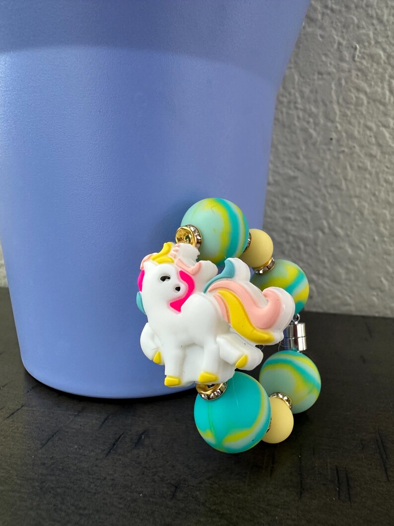 K&ouml;nnte beinhalten: Ein wei&szlig;er Einhorn-Anh&auml;nger mit rosa, gelben und blauen Akzenten ist an einem Perlenarmband befestigt. Das Armband hat bunte, marmorierte Perlen in Blau-, Gelb- und Gr&uuml;nt&ouml;nen. Der Anh&auml;nger und das Armband sind vor einem hellvioletten Hintergrund platziert.