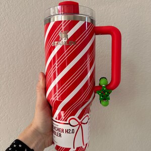 Pode incluir: Caneca Stanley Quencher H2.0 listrada em vermelho e branco com al&ccedil;a e tampa vermelhas. A caneca tem um canudo transparente e um pingente de &aacute;rvore de Natal verde preso &agrave; al&ccedil;a.