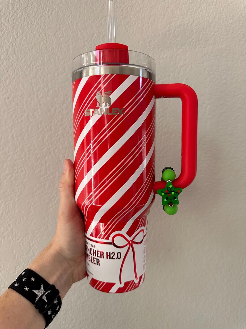 Pode incluir: Caneca Stanley Quencher H2.0 listrada em vermelho e branco com al&ccedil;a e tampa vermelhas. A caneca tem um canudo transparente e um pingente de &aacute;rvore de Natal verde preso &agrave; al&ccedil;a.