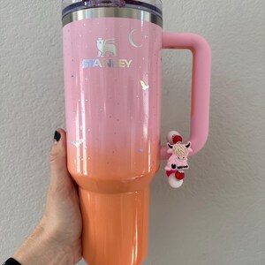 May include: A Stanley tumbler in a gradient of pink and orange, with a pink handle. The tumbler features the Stanley logo, stars, and a crescent moon design. A pink cow charm is attached to the handle.