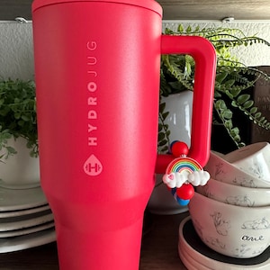 May include: A red HydroJug water bottle with a handle and a rainbow charm. The bottle has the HydroJug logo and text printed on the side. The handle has a charm with a rainbow and clouds.