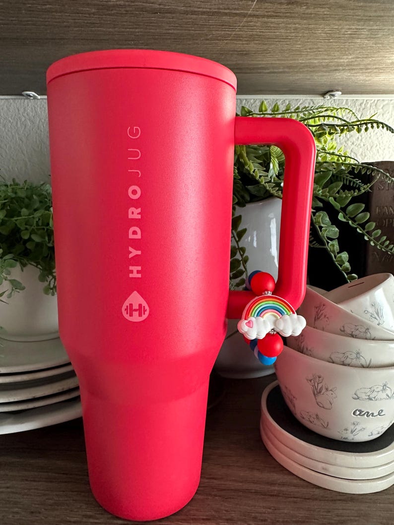 May include: A red HydroJug water bottle with a handle and a rainbow charm. The bottle has the HydroJug logo and text printed on the side. The handle has a charm with a rainbow and clouds.