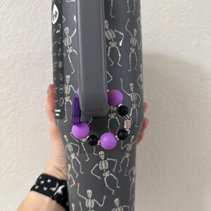 Puede incluir: Un vaso gris con asa, decorado con dise&ntilde;os de esqueletos plateados bailando. Un accesorio de cuentas moradas y negras cuelga del asa. El vaso lo sostiene una persona.